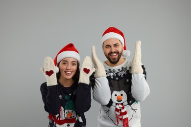 Photo of Young couple in Christmas sweaters, Santa hats and knitted mittens on grey background