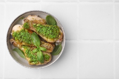Delicious fried chicken drumsticks with pesto sauce and basil in bowl on white tiled table, top view. Space for text