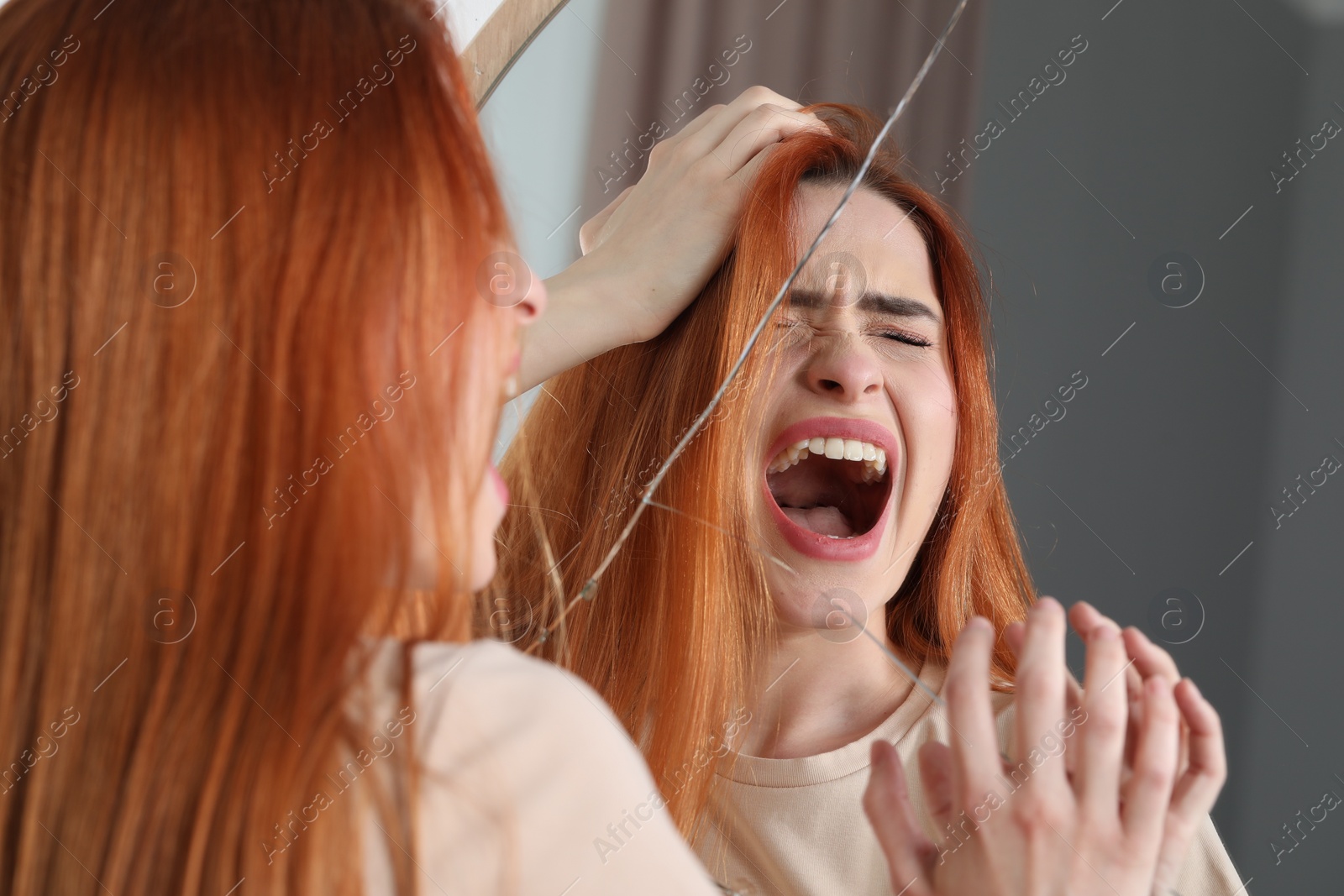 Photo of Mental problems. Young woman screaming near broken mirror indoors