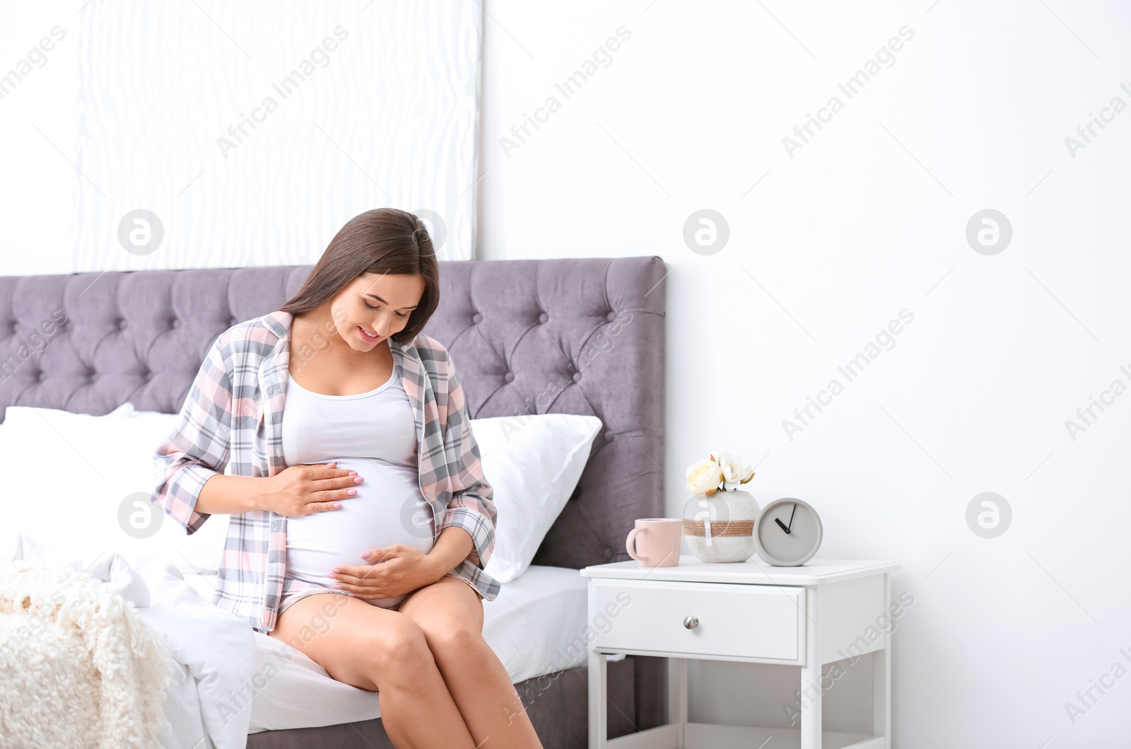 Photo of Young beautiful pregnant woman sitting on bed and touching her belly at home