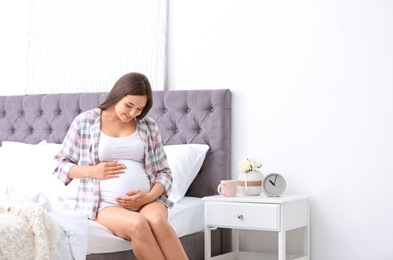 Young beautiful pregnant woman sitting on bed and touching her belly at home