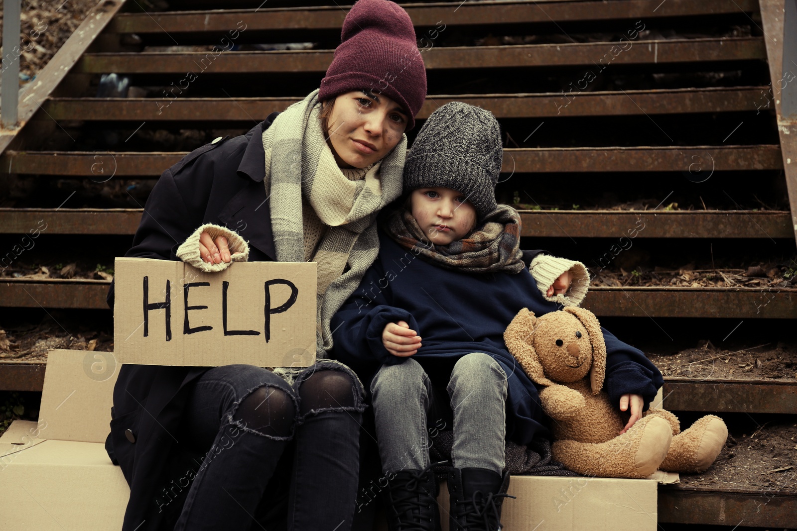 Photo of Poor mother and daughter with HELP sign sitting on stairs outdoors