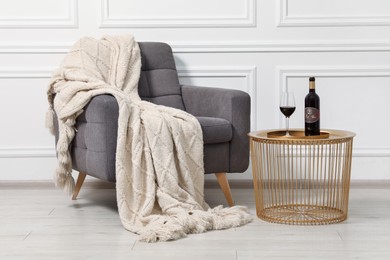 Photo of Comfortable armchair, blanket and wine on side table near white wall. Interior design