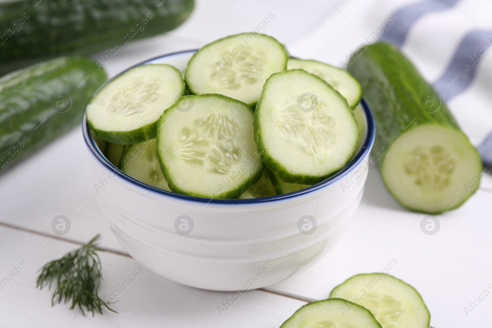 Photo of Cut cucumber in bowl, fresh vegetables and dill on white wooden table, closeup