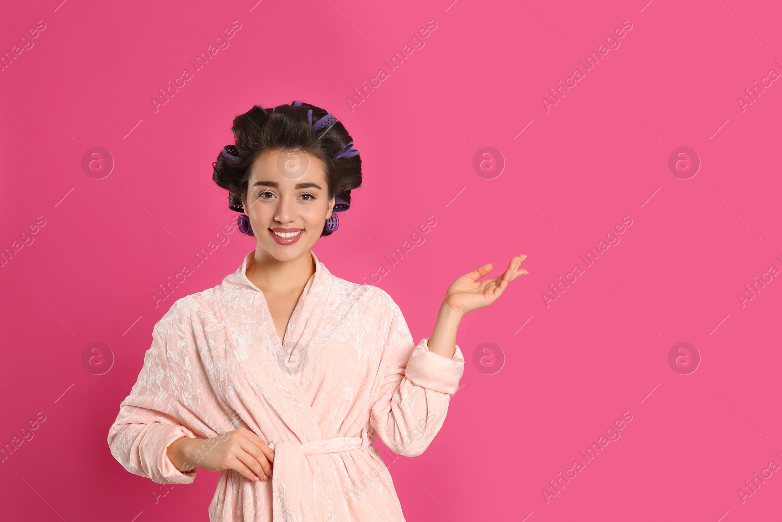 Photo of Happy young woman in bathrobe with hair curlers on pink background, space for text