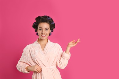 Photo of Happy young woman in bathrobe with hair curlers on pink background, space for text