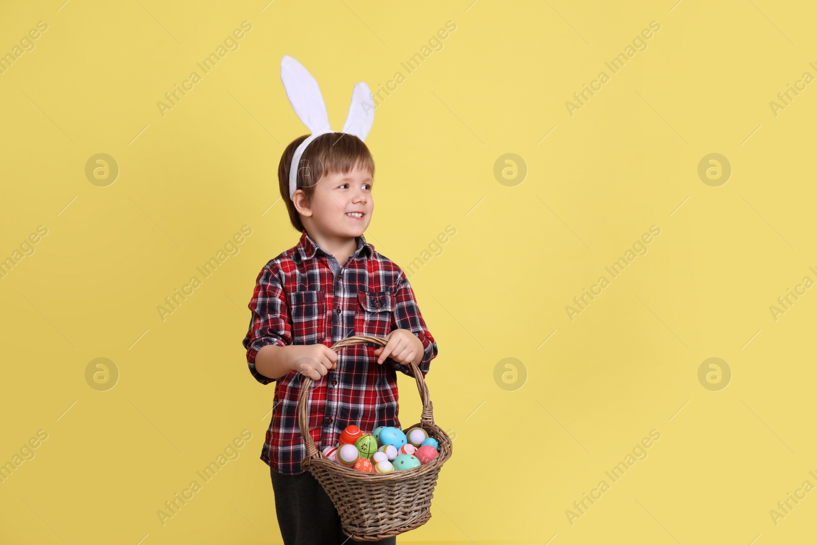 Photo of Cute little boy wearing bunny ears with basket full of dyed Easter eggs on yellow background