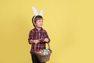 Photo of Cute little boy wearing bunny ears with basket full of dyed Easter eggs on yellow background