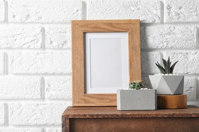 Photo of Blank frame and succulent plants on wooden cabinet near white brick wall, space for design. Home decor