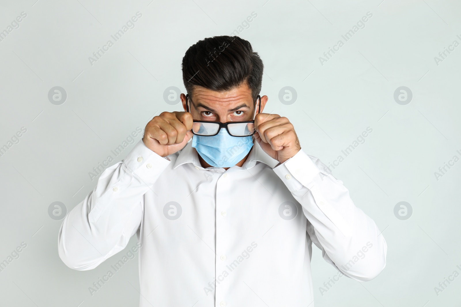 Photo of Man wiping foggy glasses caused by wearing medical mask on light background