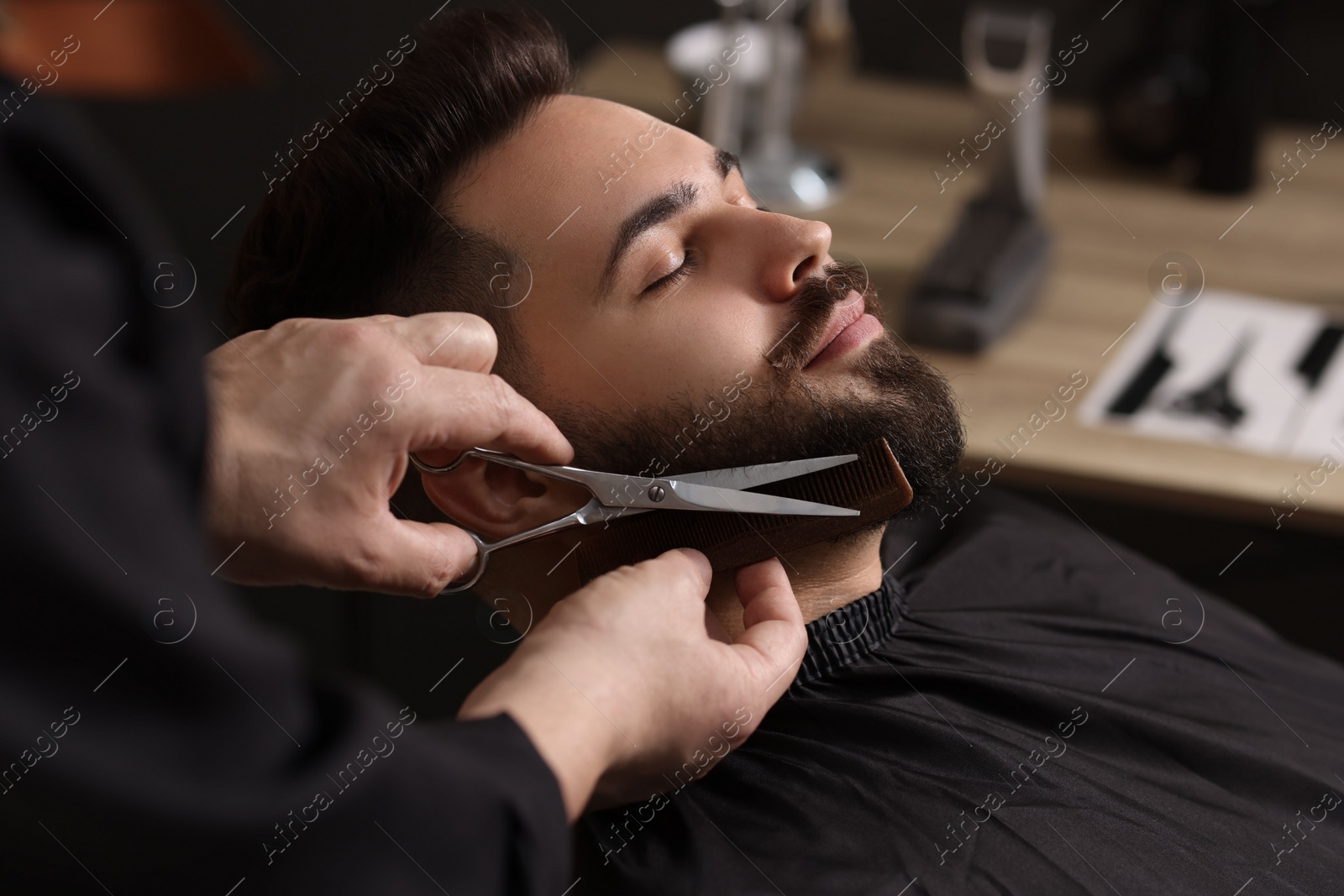 Photo of Professional barber trimming client's beard with scissors in barbershop, closeup