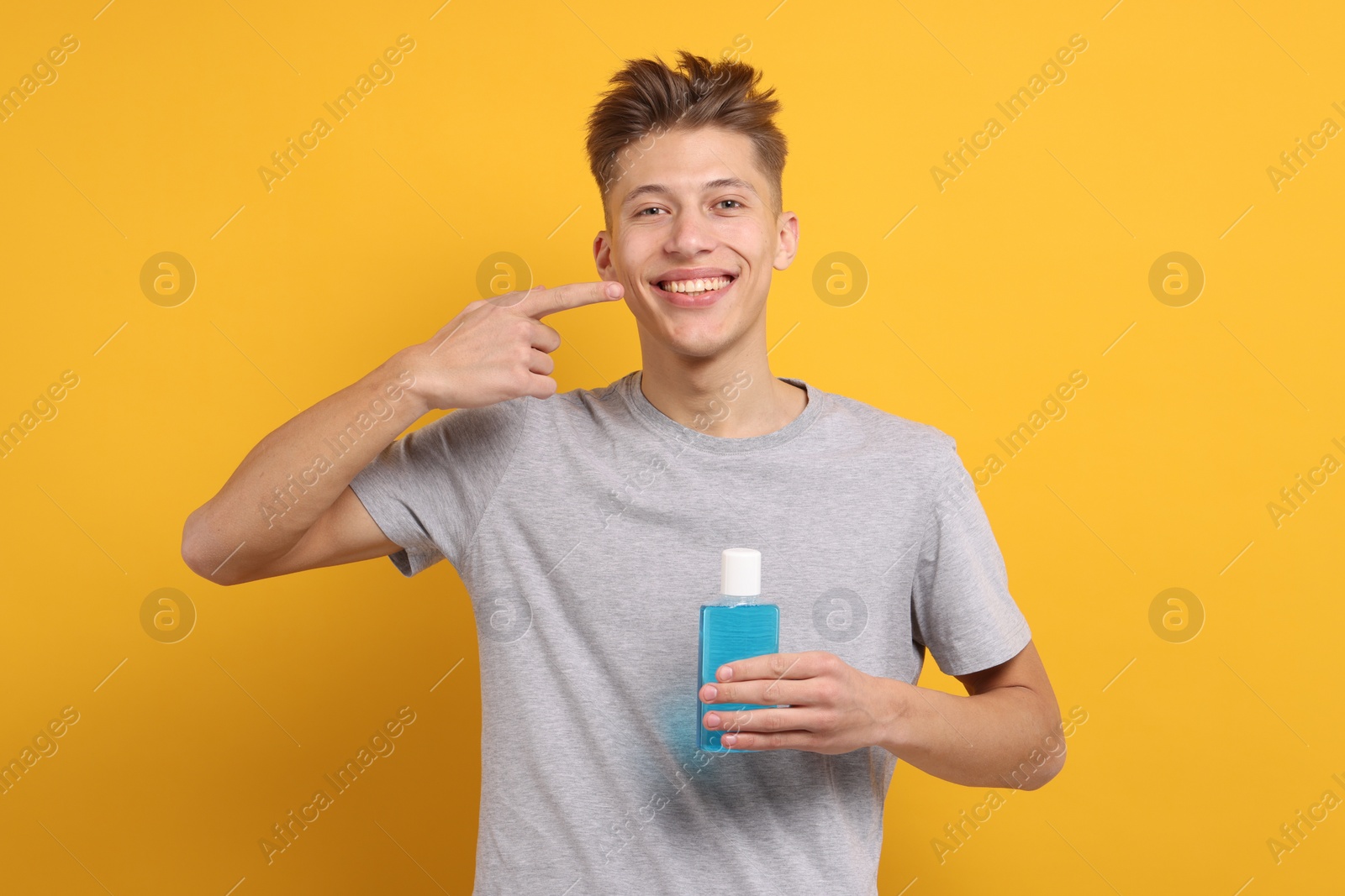 Photo of Young man with mouthwash pointing at his healthy teeth on yellow background