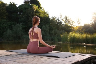 Woman practicing Padmasana on yoga mat on wooden pier near pond, back view. Lotus pose