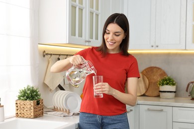 Photo of Young woman pouring water into glass from jug in kitchen