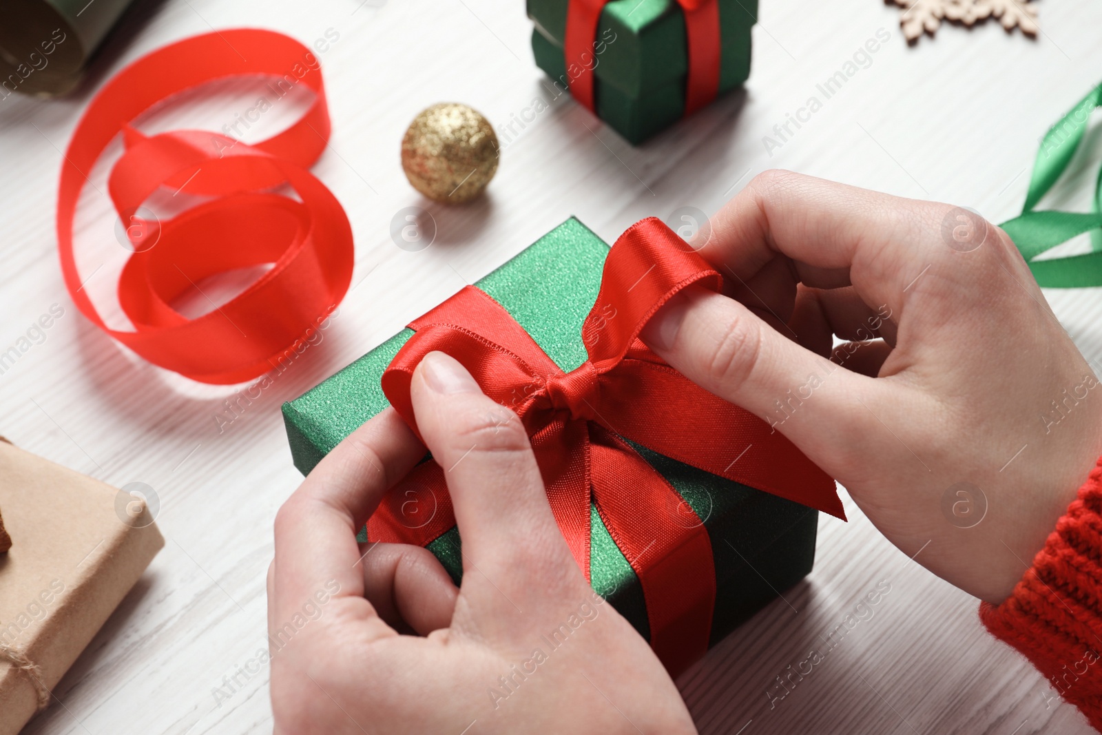 Photo of Christmas present. Woman tying ribbon bow on gift box at white wooden table, closeup