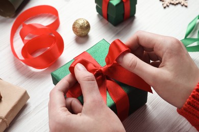 Photo of Christmas present. Woman tying ribbon bow on gift box at white wooden table, closeup