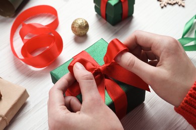 Christmas present. Woman tying ribbon bow on gift box at white wooden table, closeup