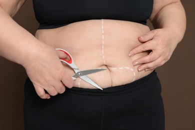 Photo of Obese woman with scissors and marks on body against brown background, closeup. Weight loss surgery