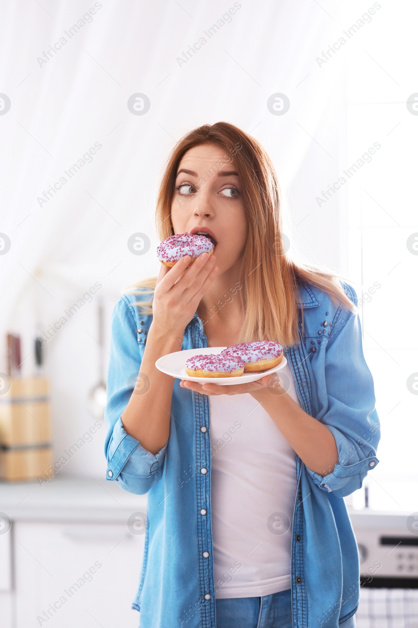 Photo of Emotional young woman eating donut in kitchen. Failed diet