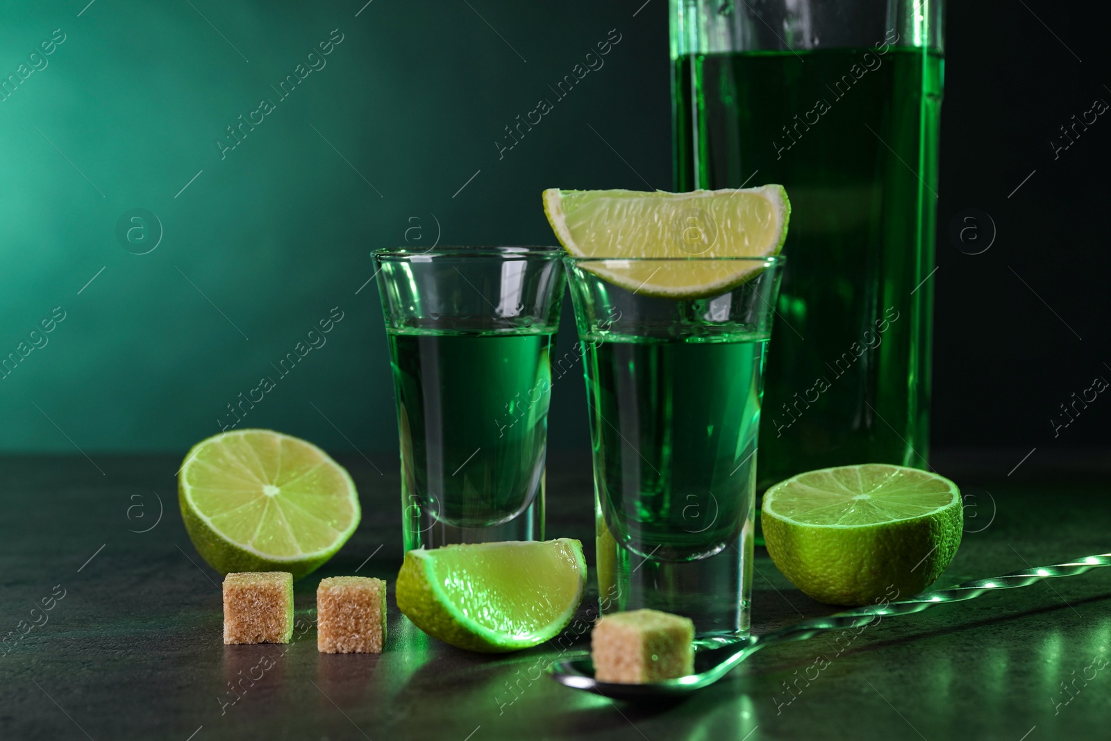 Photo of Absinthe in shot glasses, spoon, brown sugar cubes and lime on gray textured table against green background, space for text. Alcoholic drink