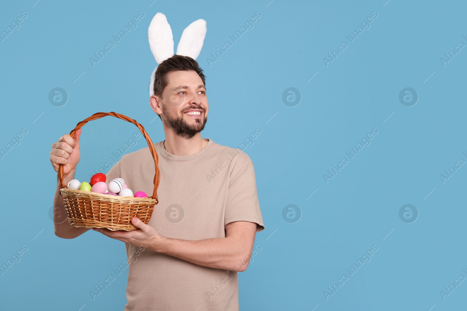 Photo of Happy man in bunny ears headband holding wicker basket with painted Easter eggs on turquoise background. Space for text