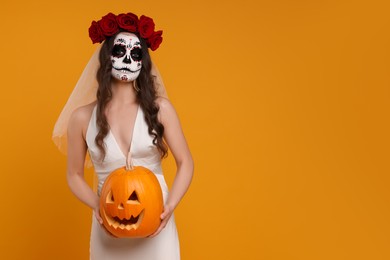 Photo of Young woman in scary bride costume with sugar skull makeup, flower crown and carved pumpkin on orange background, space for text. Halloween celebration
