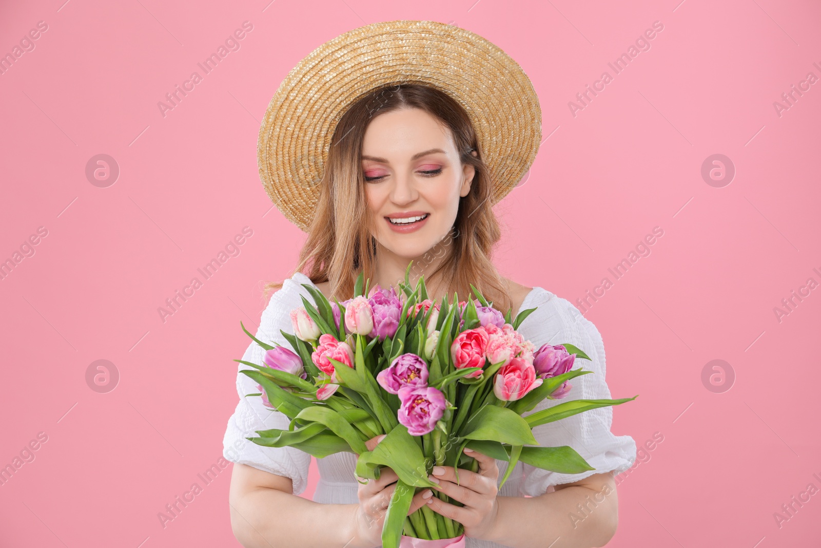 Photo of Happy young woman in straw hat holding bouquet of beautiful tulips on pink background