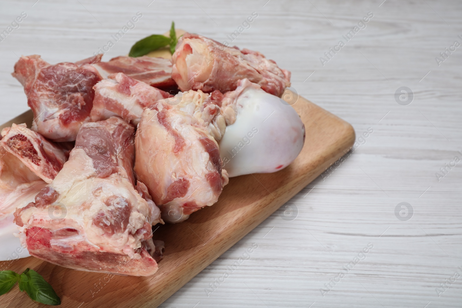 Photo of Cutting board with raw chopped meaty bones and basil on white wooden table, closeup. Space for text