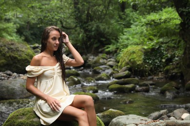 Photo of Beautiful young woman sitting on rock near mountain river in forest. Space for text