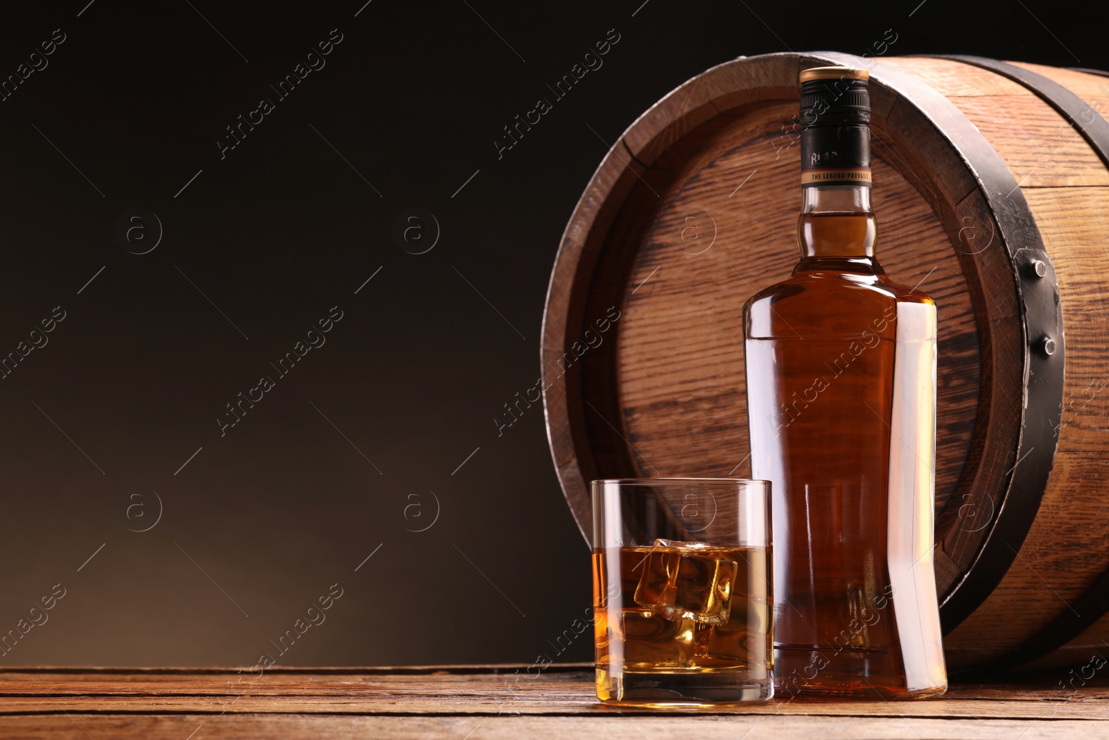 Photo of Whiskey with ice cubes in glass, bottle and barrel on wooden table against black background, space for text