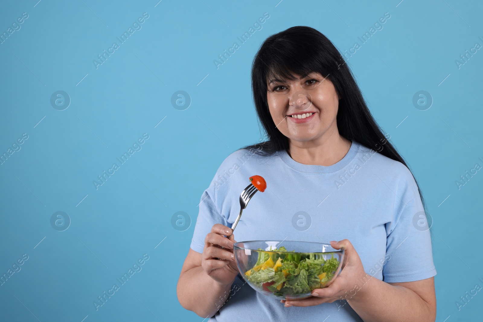 Photo of Beautiful overweight woman eating salad on light blue background, space for text. Healthy diet