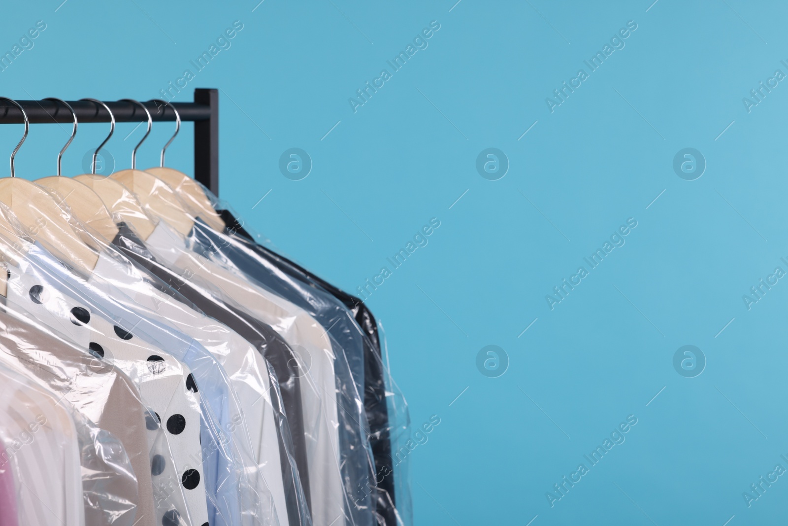 Photo of Dry-cleaning service. Many different clothes in plastic bags hanging on rack against light blue background, space for text