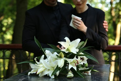 Couple near granite tombstone with white lilies at cemetery outdoors, selective focus. Funeral ceremony