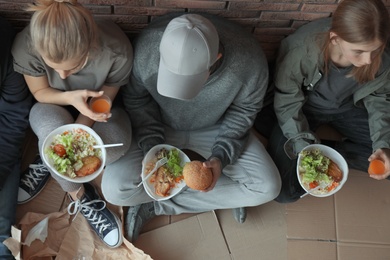 Photo of Poor people with plates of food sitting at wall indoors, view from above