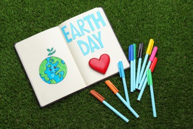 Photo of Notebook with words Earth Day, planet drawing and markers on green grass, flat lay