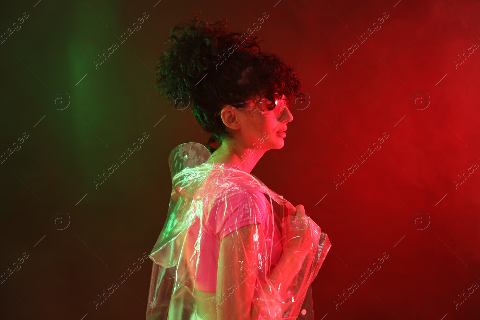 Photo of Beautiful young woman in transparent coat and sunglasses posing on color background in neon lights