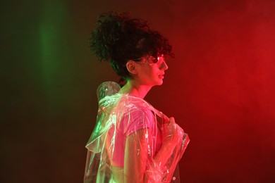 Photo of Beautiful young woman in transparent coat and sunglasses posing on color background in neon lights