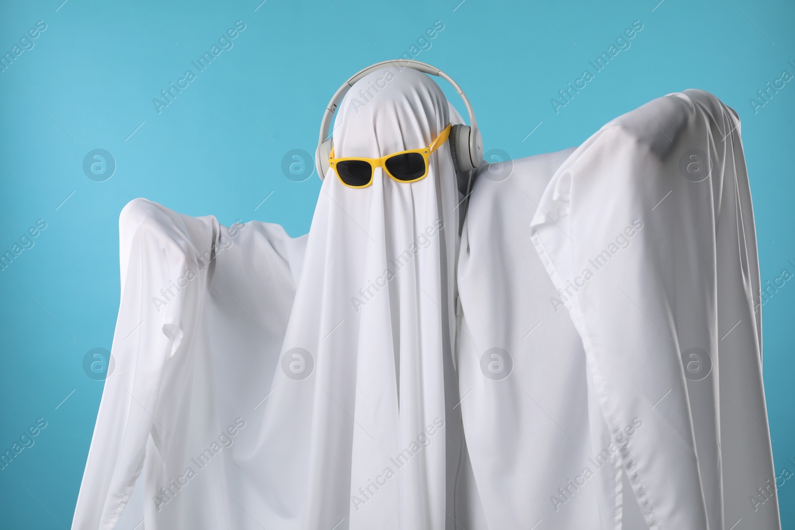 Photo of Stylish ghost. Person covered with white sheet in sunglasses and headphones on light blue background