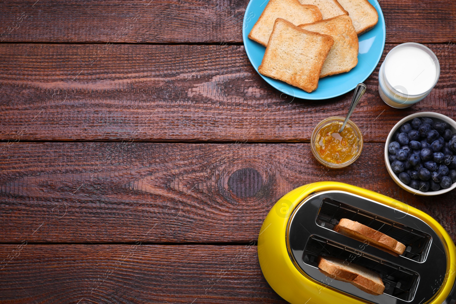 Photo of Yellow toaster with roasted bread, glass of milk, blueberries and jam on wooden table, flat lay. Space for text