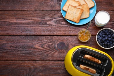 Photo of Yellow toaster with roasted bread, glass of milk, blueberries and jam on wooden table, flat lay. Space for text