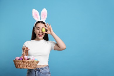 Happy woman in bunny ears headband holding wicker basket of painted Easter eggs on turquoise background. Space for text