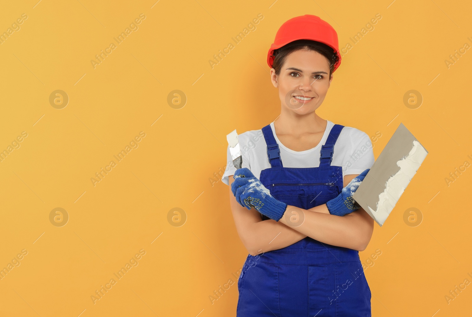 Photo of Professional worker with putty knives in hard hat on orange background, space for text