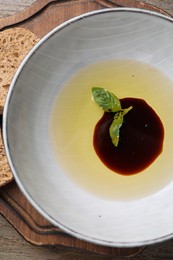 Photo of Bowl of balsamic vinegar with oil and basil served with bread slices on wooden board, closeup