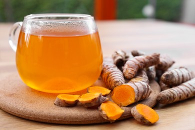 Photo of Glass cup of hot tea and fresh turmeric roots on wooden table, closeup
