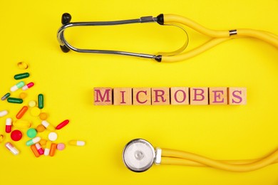 Photo of Word Microbes made with wooden cubes, pills and stethoscope on yellow background, top view