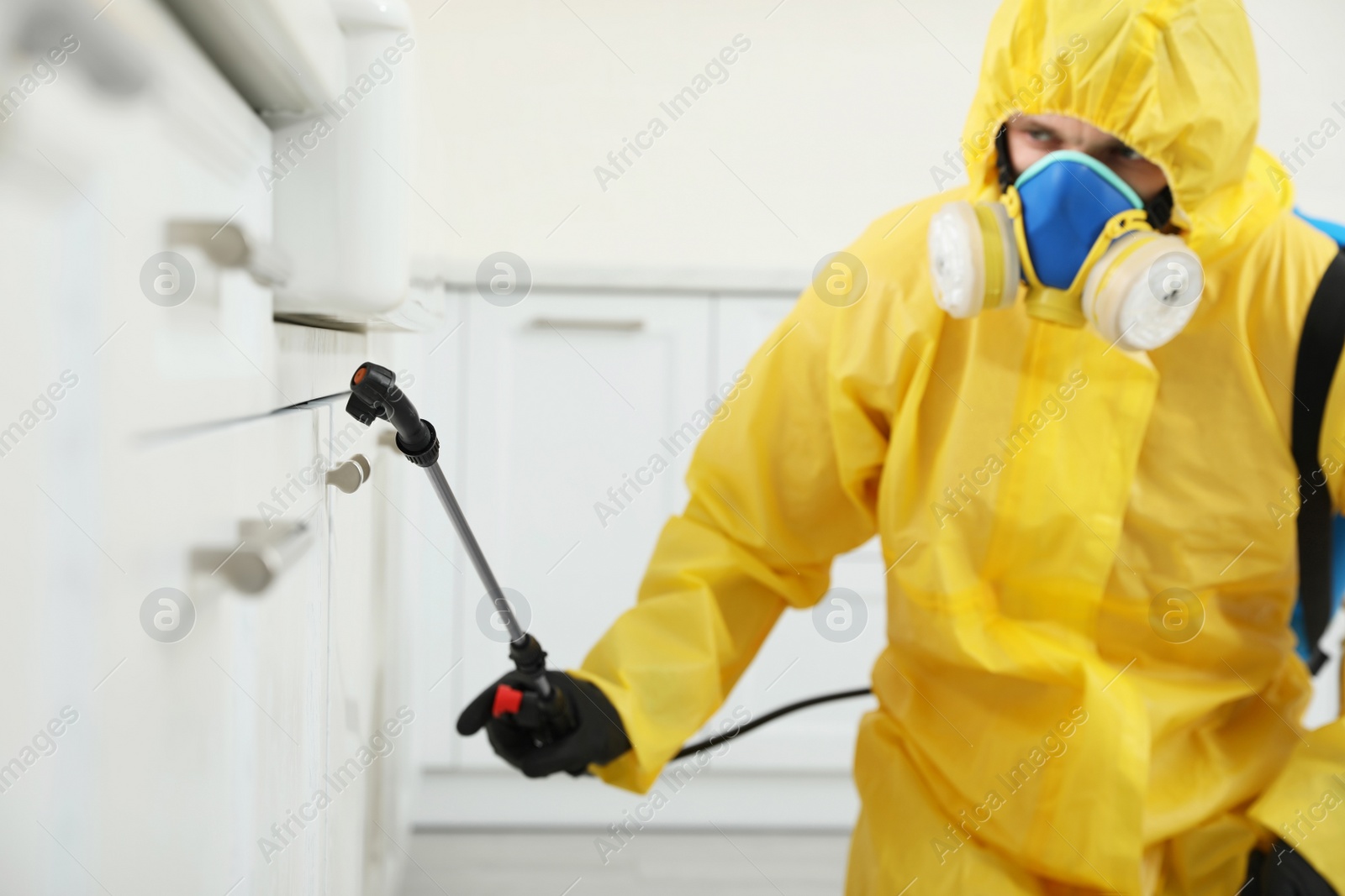 Photo of Pest control worker in protective suit spraying insecticide on furniture indoors