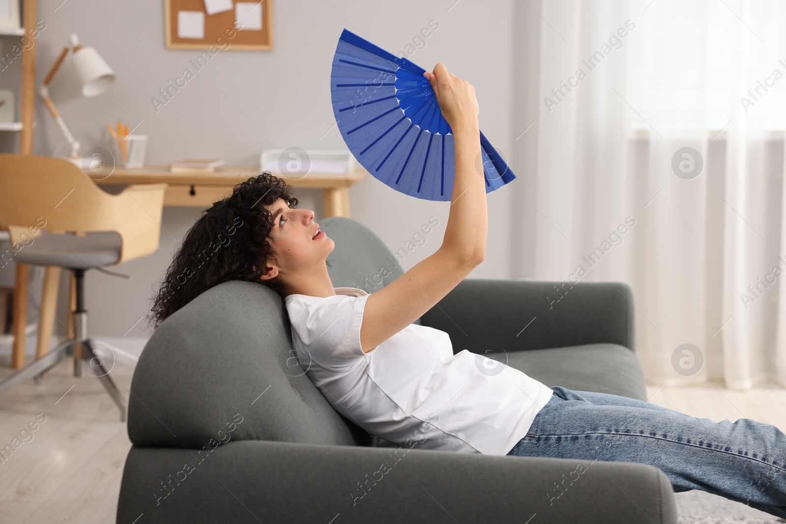 Photo of Young woman waving blue hand fan to cool herself on sofa at home