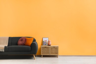 Stylish grey sofa with colorful pillows and wooden cabinet near pale orange wall indoors, space for text. Interior design