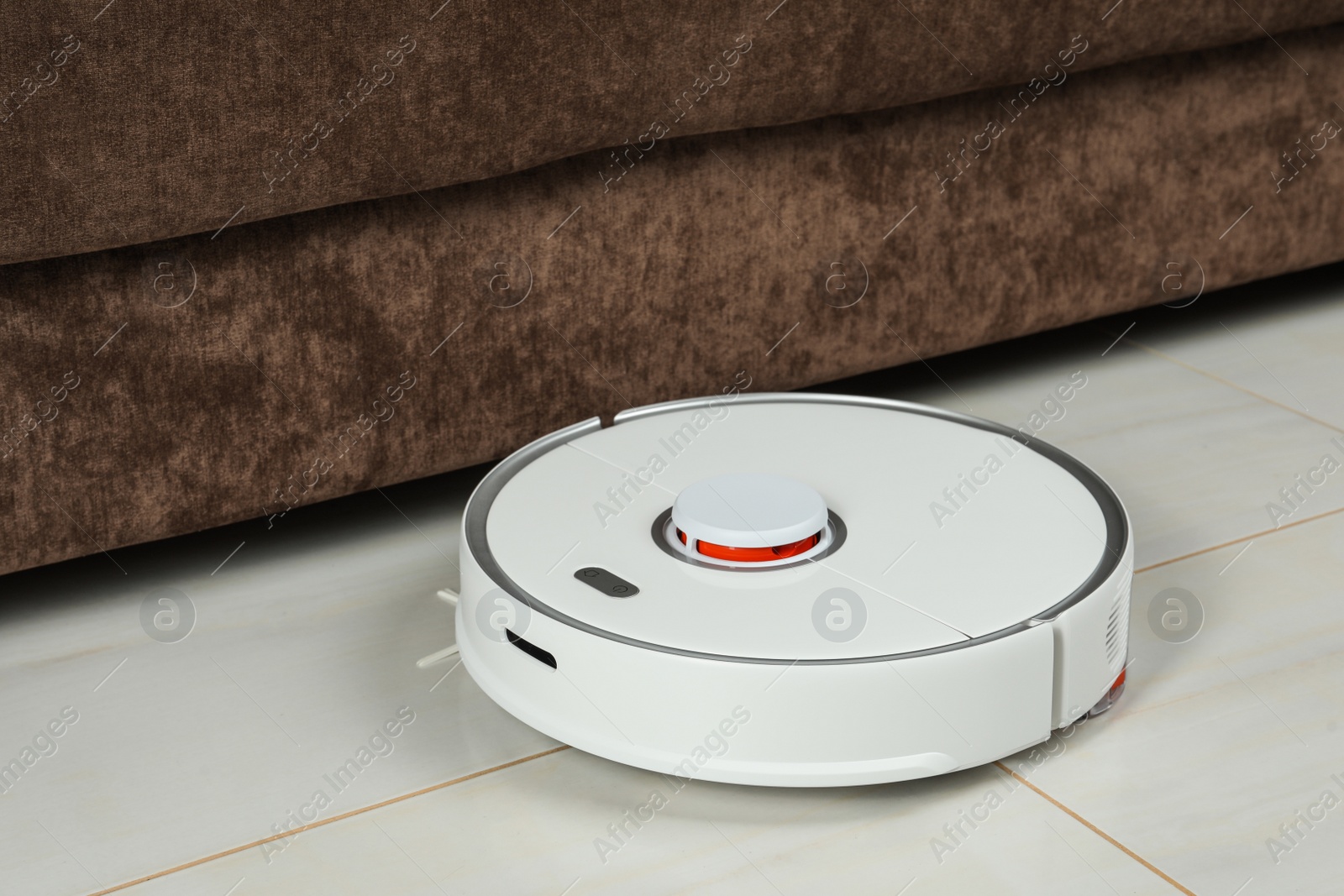 Photo of Robotic vacuum cleaner on white tiled floor near sofa indoors