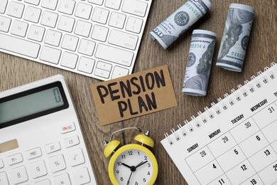 Photo of Card with words Pension Plan, banknotes, calculator and alarm clock on wooden office table, flat lay